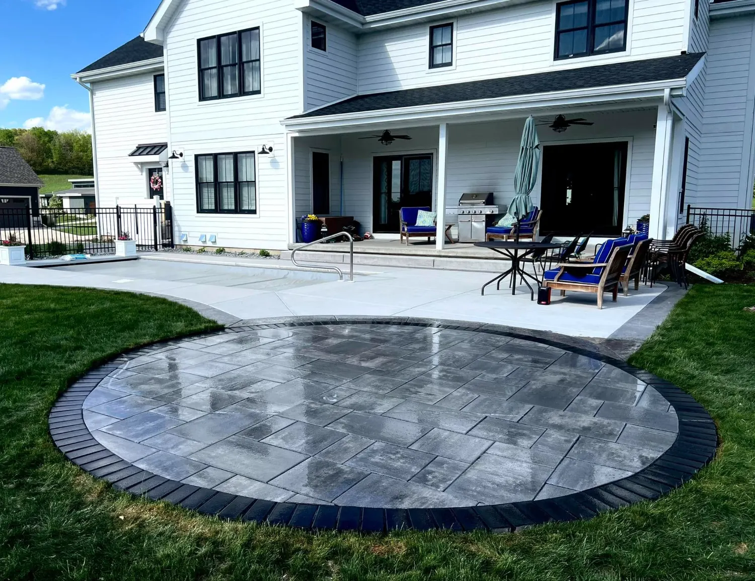 Circular grey paver patio set into a broom-finish concrete patio behind a white farmhouse