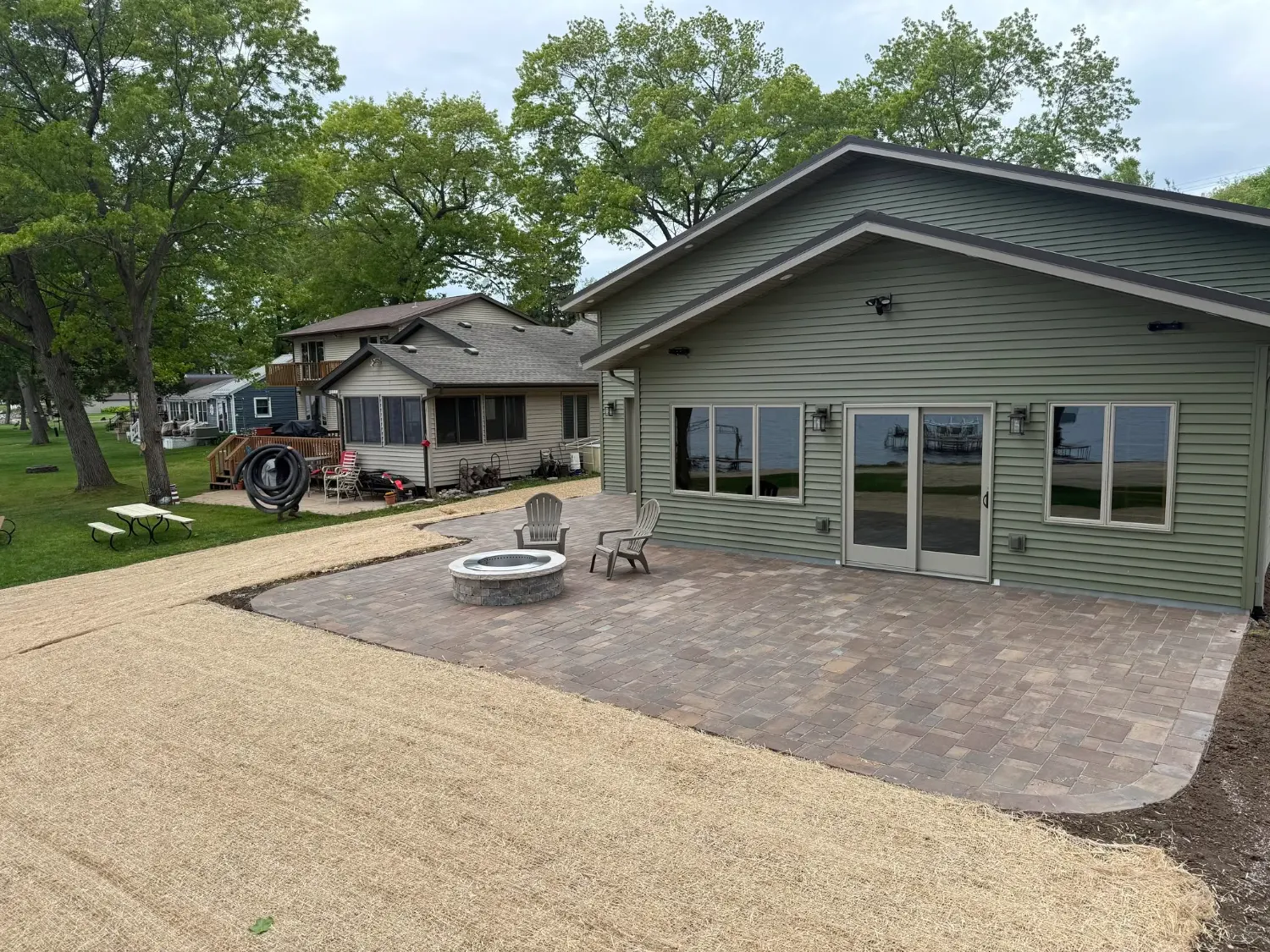 Tumbled paver patio with a round fire pit behind a green lakeside house