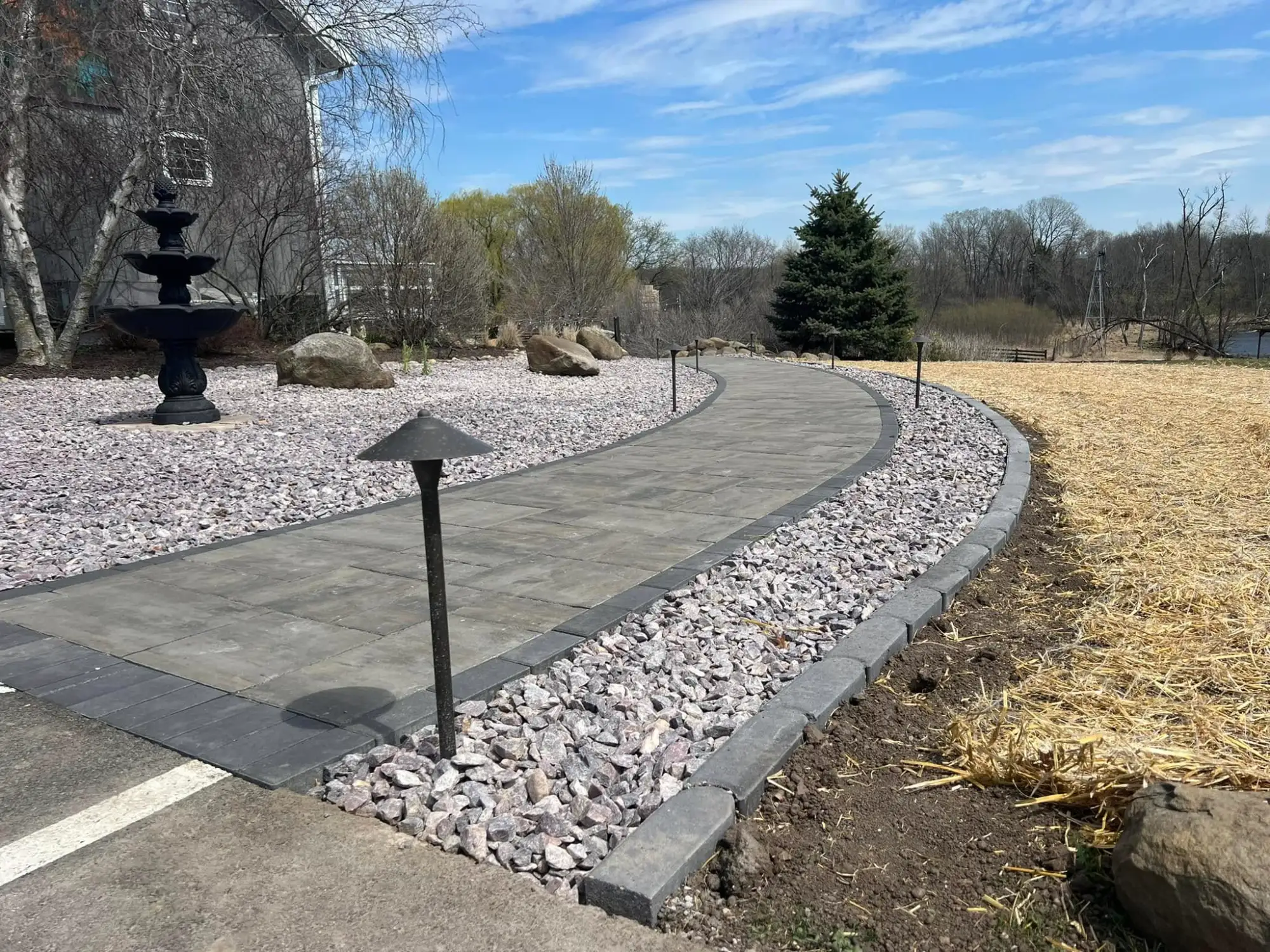 A curved paver walkway edged in charcoal stone, lit by low landscape lights, curving toward the water on a spring afternoon