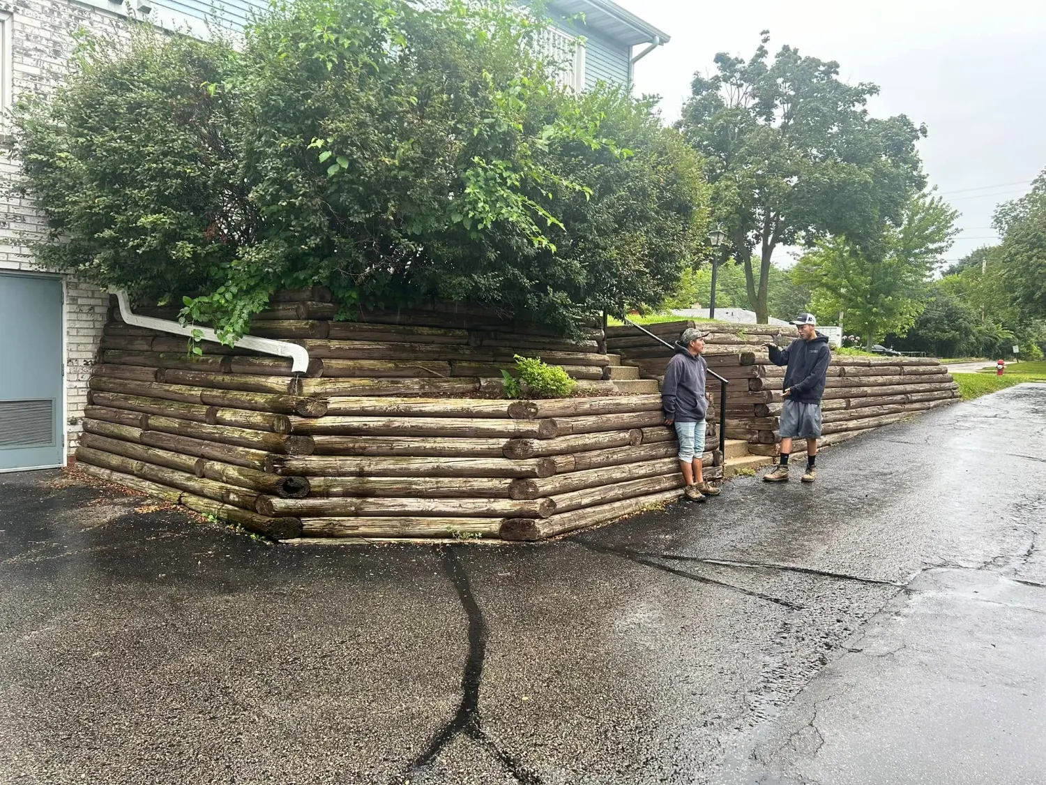 Two crew members inspecting the failing stacked-timber retaining wall on a wet morning